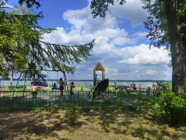 Nad jeziorem ogrodzony plac zabaw ze sztuczną nawierzchnią.