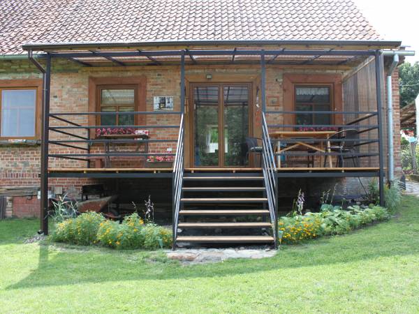 Duży, zadaszony taras z modrzewiową deską. Na tarasie dębowy stół i krzesła. Schody w kierunku