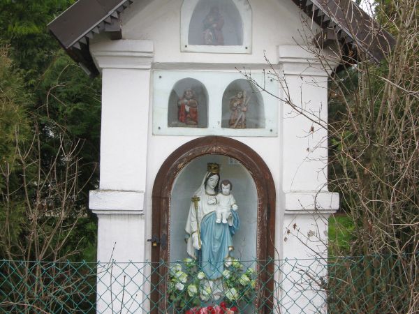 kapliczka przydrożna, zabytkowa, z figurą Matki Boskiej z Dzieciątkiem - w niszy głównej - za szkłem oraz wyżej w płycinach za taflami z pleksy: miniaturowe rze