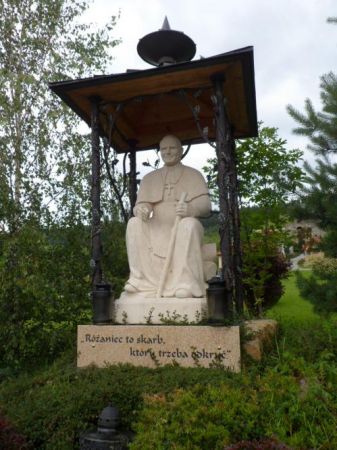 figura św. Jana Pawła II siedzącego na pniaczku z różańcem w ręku i uśmiechającego się do gór, które tak kochał.