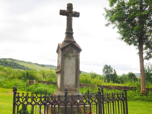 nagrobek kamienny - krzyż na cokole, kunsztownie wykonany, otoczony kutym płotkiem, dalej widoczny murek z kamieni polnych.