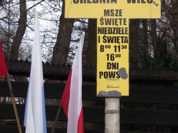 na planie krzyża : kościelna / parafialna tablica informacyjna, dalej fragment stylowego ogrodzenia.