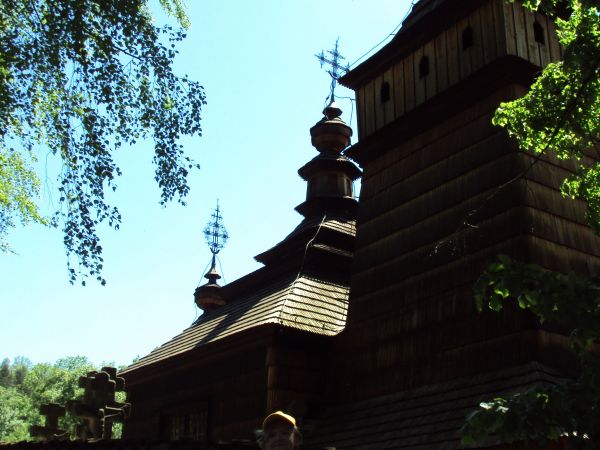 cerkiew otoczona pozornym drewnianym parkanem na kamiennej podmurówce, w cieniu wiekowych drzew.