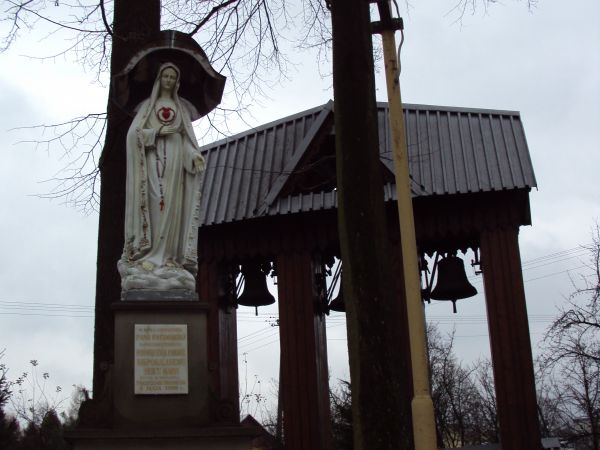 figura Matki Boskiej Fatimskiej na postumencie, dalej: stylowa parawanowa dzwonnica. Za dziedzińcem kościelnym droga do uzdrowiska w sąsiedztwie wzgórza Cyrenaj