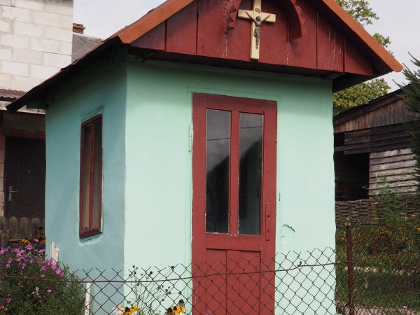 kapliczka domkowa murowana, nakryta dwuspadowym dachem pokrytym blachą, na nim blaszana kopułka zwieńczona metalowym krzyżem z Wizerunkiem Ukrzyżowanego nakryty