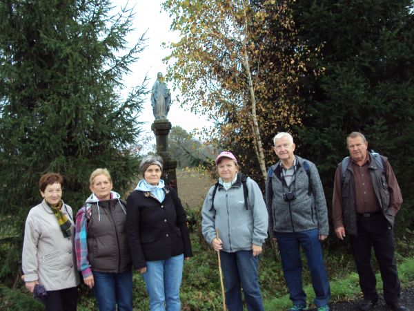 przed / obok: przydrożna kapliczka słupowa z figurą Matki Bożej na cokole. Figura przedstawia Matkę Boską, która stoi na kuli ziemskiej, którą opasuje wąż. Dwuk