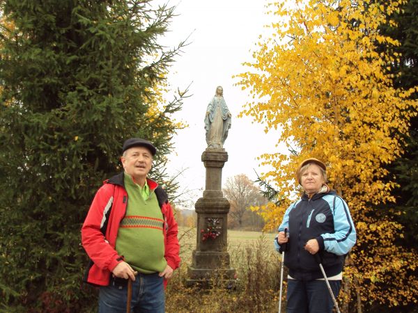 przydrożna kapliczka słupowa z figurą  Matki Bożej na cokole, dwukondygnacyjna, na płycinie poniżej: przytwierdzona postać Chrystusa Ukrzyżowanego - na skraju w