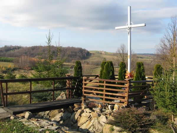przy drodze, za umocnionym kamieniami na skarpie rowem a nad nim ława / mostek : krzyż wotywny z przesłaniem / tablicą u podnóża, otoczony solidnym, drewnianym