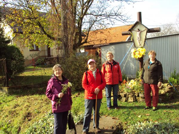 przed / obok: krzyż przydrożny w medalionie przedstawienie Chrystusa Ukrzyżowanego, otoczenie ukwiecone, zadbane, otoczone pozornym murkiem z surowego kamienia