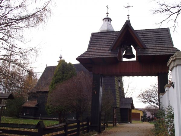 u wejścia na plac kościelny stoi stylowa, aczkolwiek współczesna dzwonnica z bramką, widoczny fragment ogrodzenia a po prawej murowanego parkanu okalającego par