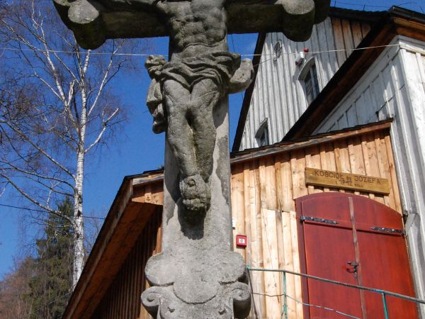 Figurka Jezusa ukrzyżowanego. W oddali drewniany Kościół - pierwotnie ewangelicki. Obecnie należy do parafii św. Józefa Oblubieńca NMP z pięknym barokowym wnętr