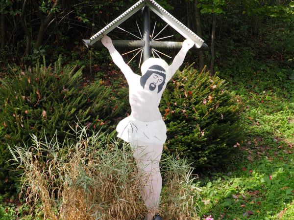 Wizerunek Pana Jezusa Ukrzyżowanego, nakryty pozornym daszkiem, na krzyżu przydrożnym, ukwiecony.