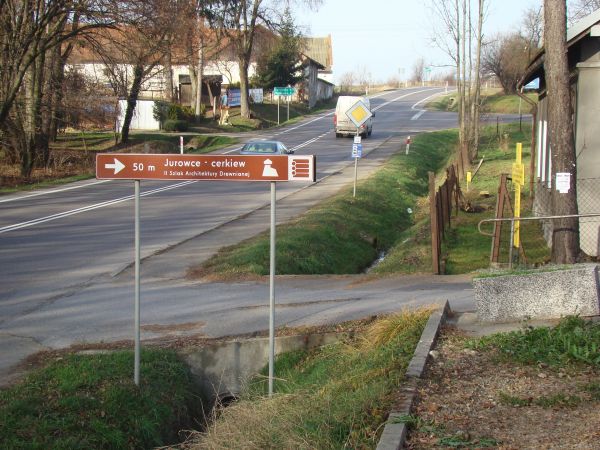 oznaczenia Szlaku Architektury Drewnianej - tu sygnalizowana cerkiew, dzwonnica i sektor cmentarny z bramą. Po lewej fragment parku dworskiego. Należy pamiętać,
