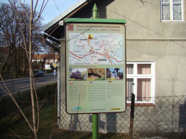 plansza "Szlaku Architektury Drewnianej" - przy kościele obok parkingu, dalej  budynek dawnej szkoły, po lewej droga Rzeszów - Brzozów - Sanok a za nią fragment