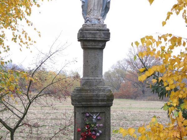 przydrożna kapliczka słupowa z figurą  Matki Bożej na cokole. Figura przedstawia Matkę Boską, która stoi na kuli ziemskiej, którą opasuje wąż. Dwukondygnacyjna,